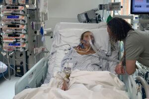 Stephanie on a ventilator lying in a hospital bed. Her boyfriend is leaning over her.